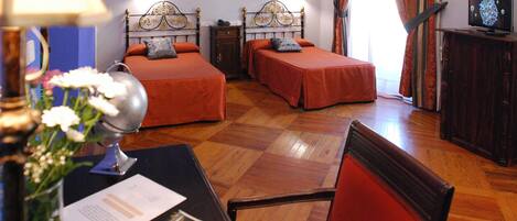 A hotel room with two single beds, a wooden floor, a desk with a lamp, and a television.