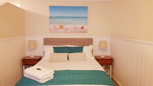Una cama bien hecha con una colcha color turquesa, dos almohadas y una toalla blanca doblada. Hay dos mesitas de noche con lámparas, y un cuadro con una escena de playa sobre la cama.