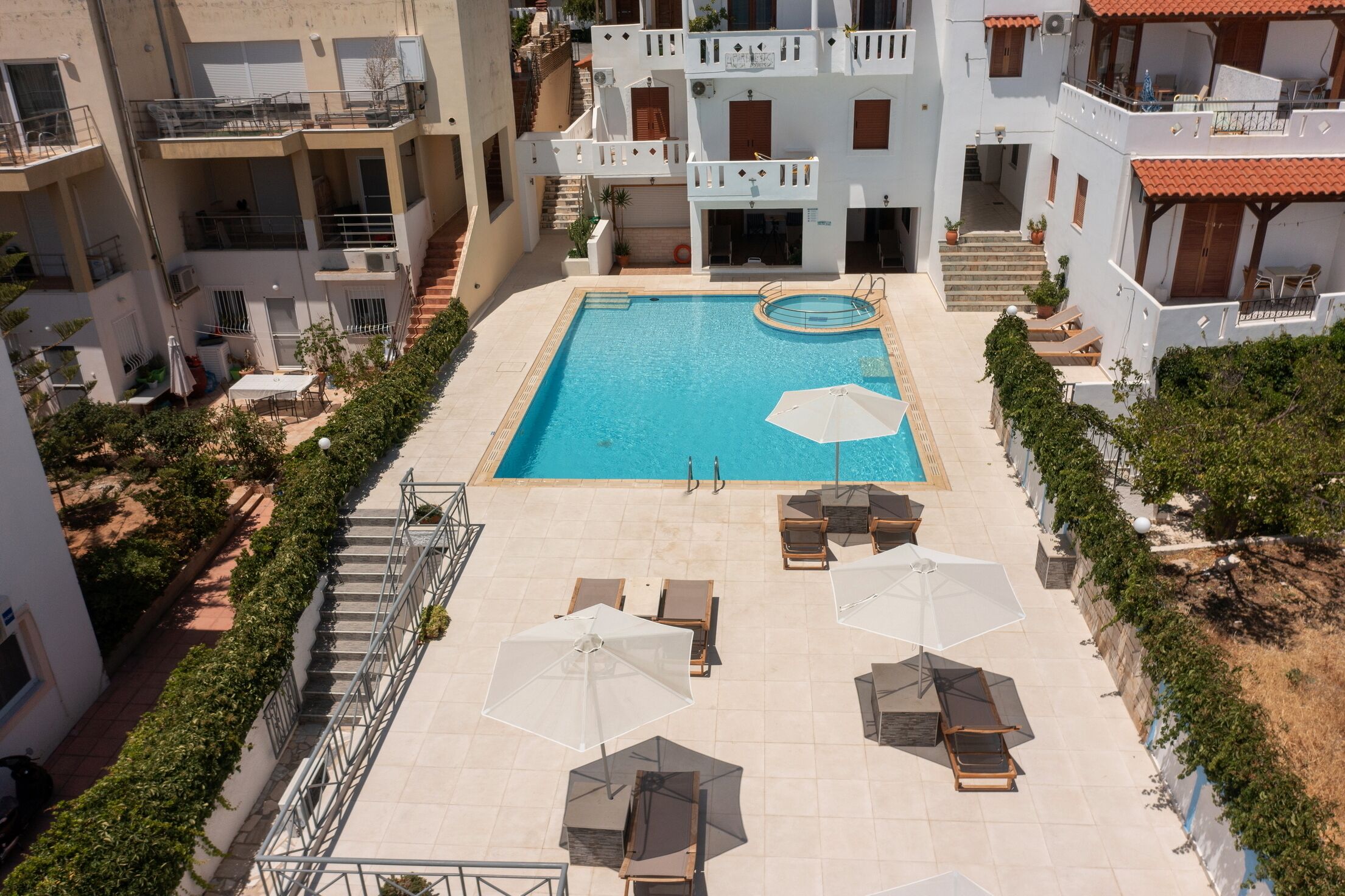 An aerial view of a hotel with a swimming pool surrounded by lounge chairs and umbrellas, with multiple balconies and staircases.