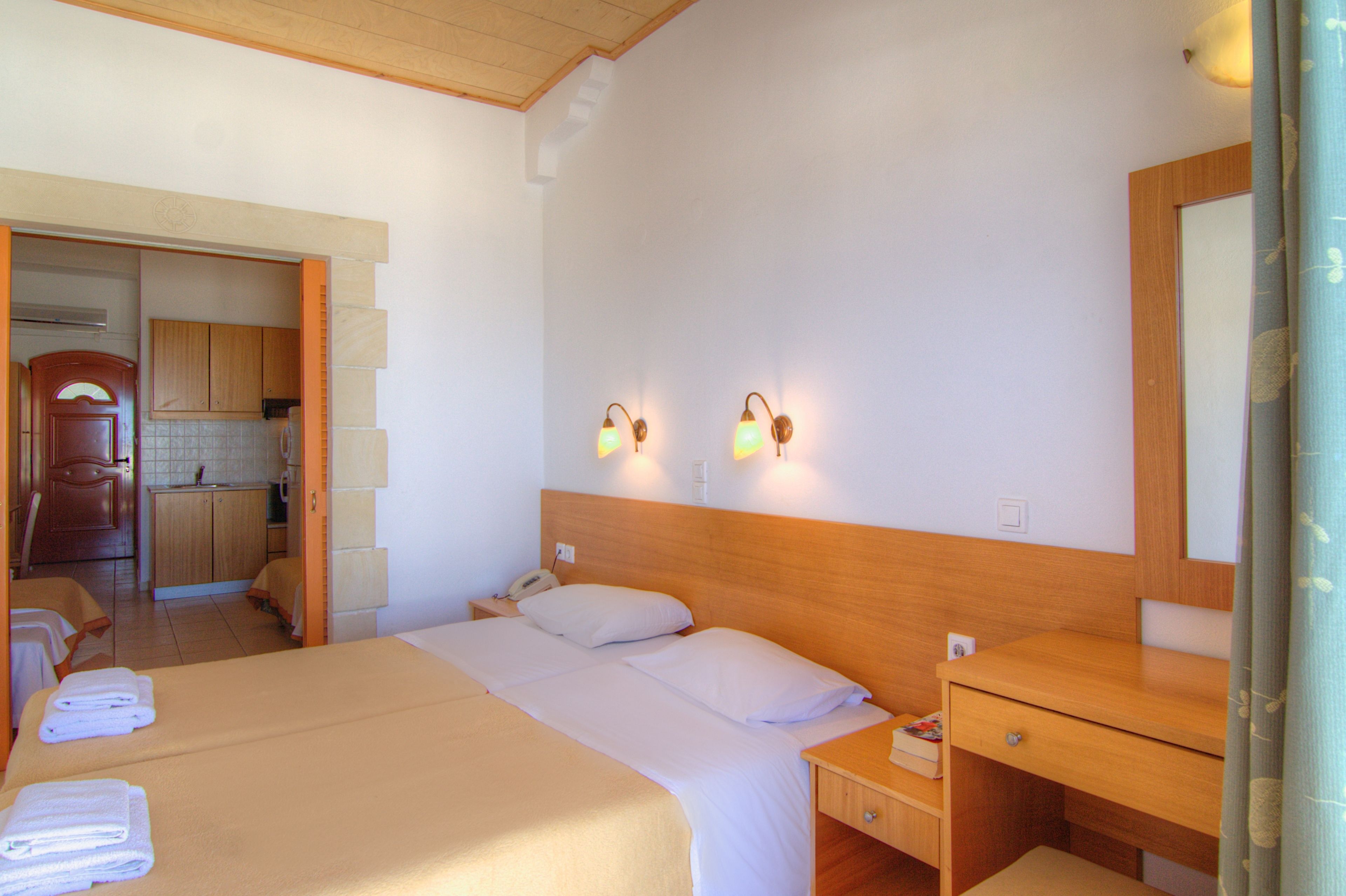 Habitación de hotel con cama, mesita de noche y cabecera de madera. Hay un escritorio pequeño con silla y un espejo. La habitación tiene una kitchenette con armarios y un lavamanos.