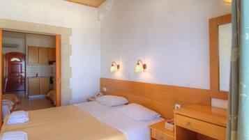 Habitación de hotel con cama, mesita de noche y cabecera de madera. Hay un escritorio pequeño con silla y un espejo. La habitación tiene una kitchenette con armarios y un lavamanos.