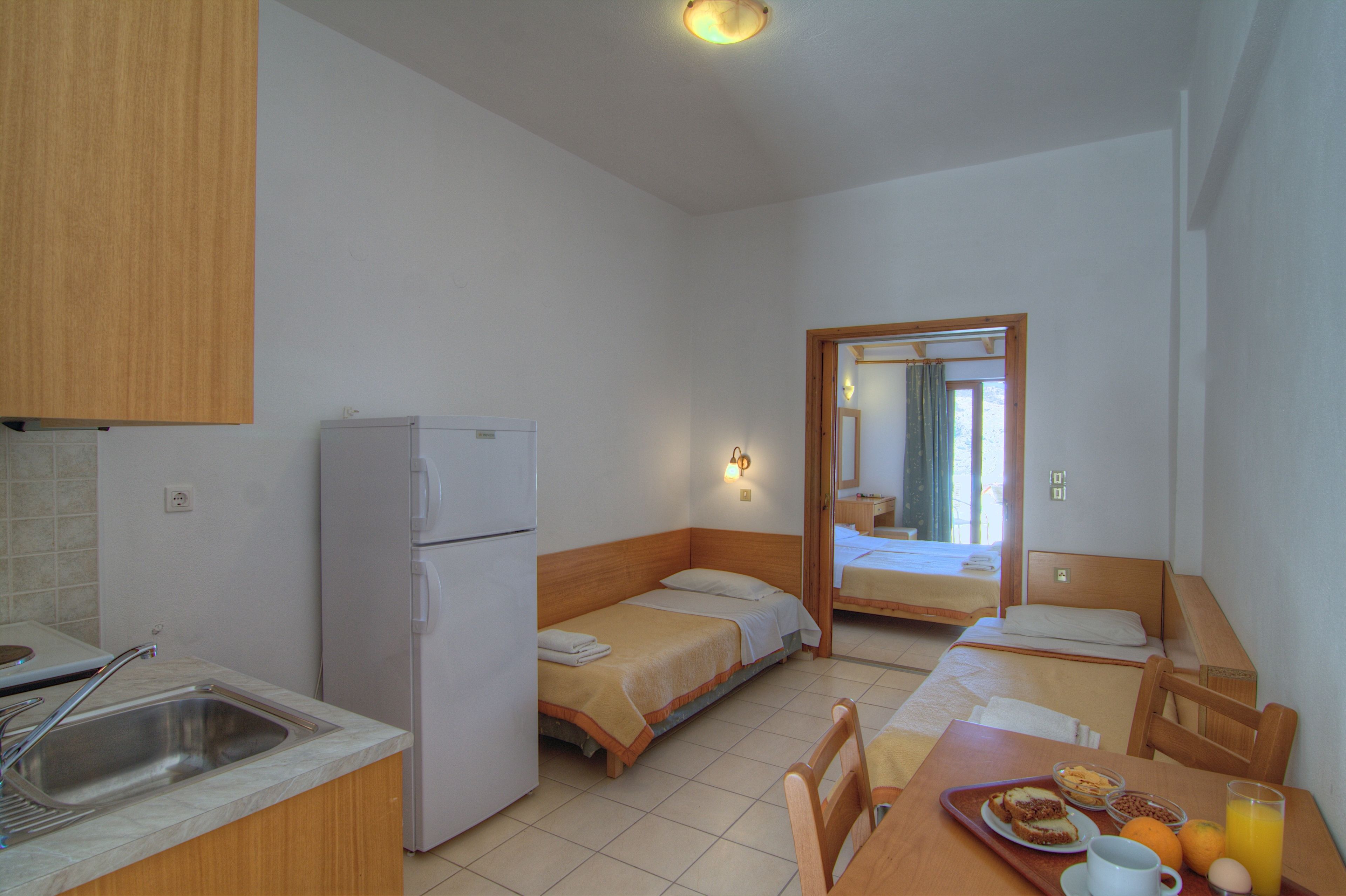 A compact hotel room with a kitchenette, two single beds, a dining table, and a small bathroom.