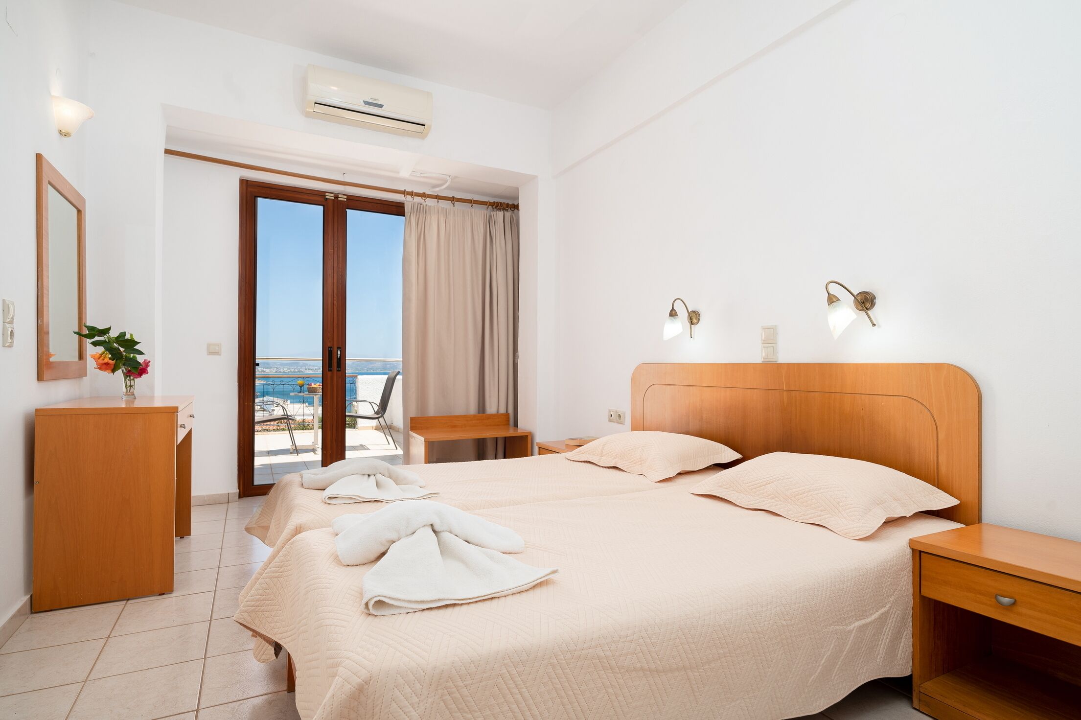 Een hotelkamer met twee bedden, een houten hoofdeinde, een nachtkastje, een bureau, een stoel, een raam met gordijnen en een airconditioning.