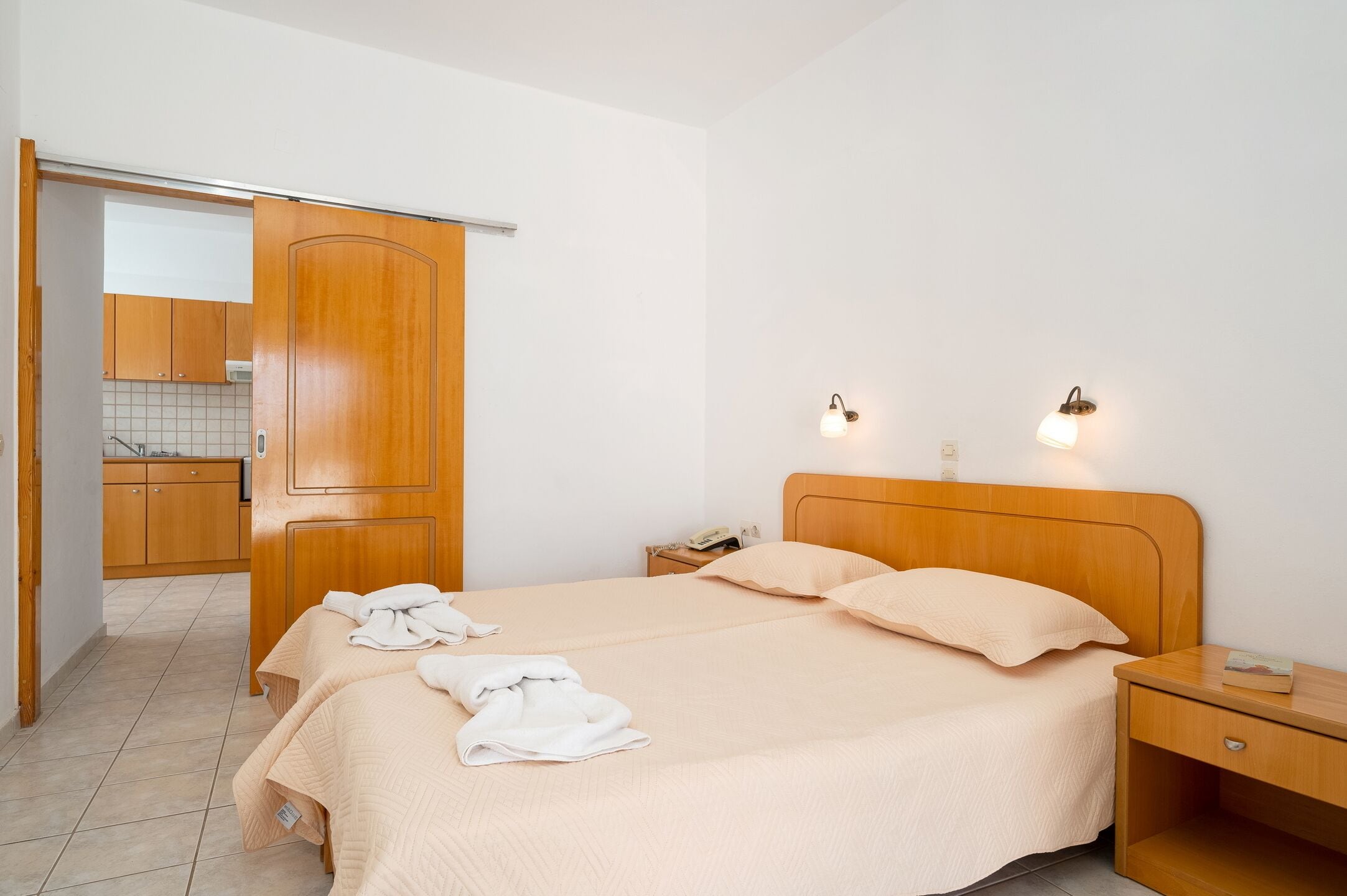 Habitación de hotel con una cama de madera, dos mesitas de noche, un armario y una cocina visible a través de una puerta abierta.