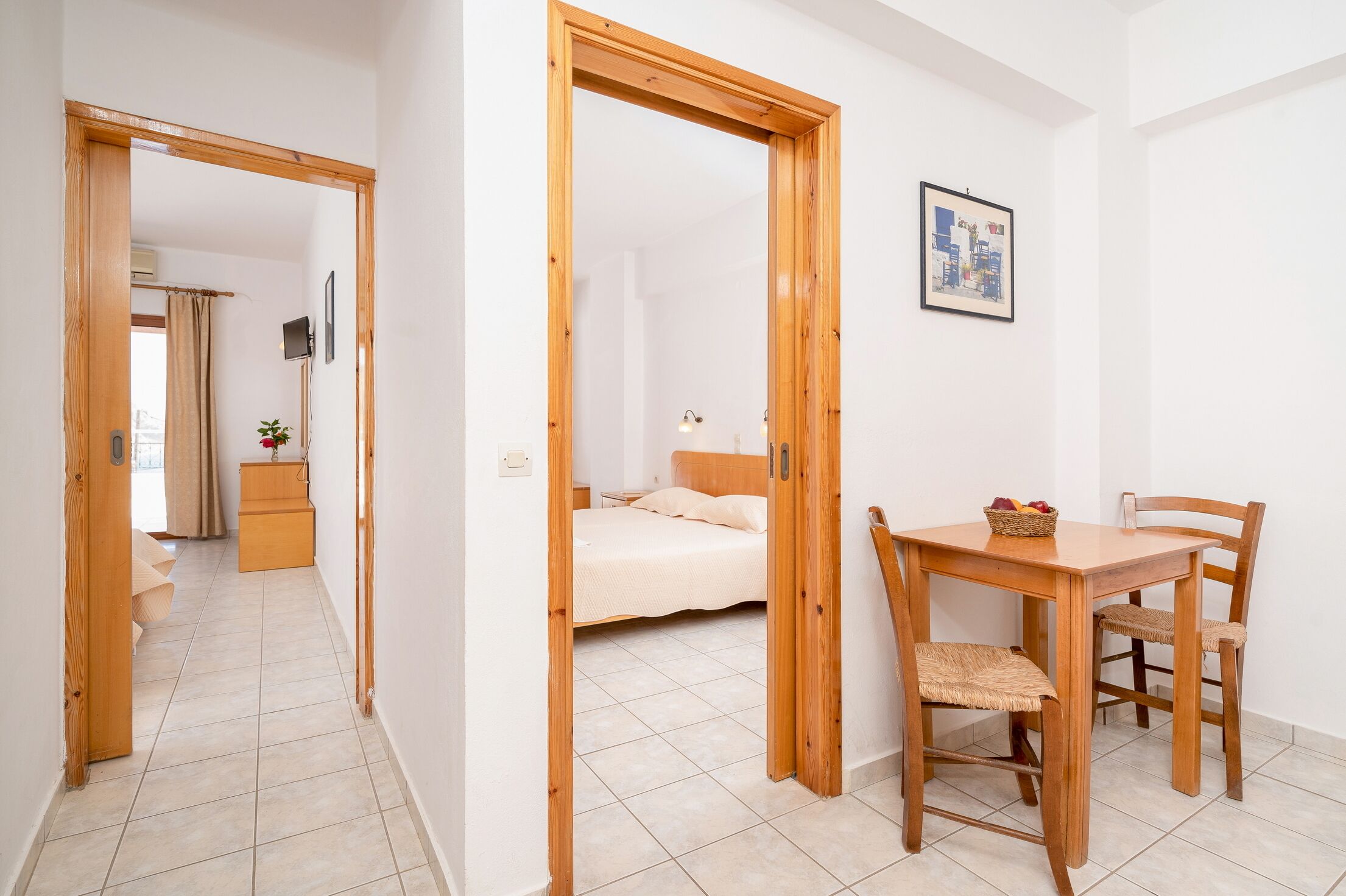 Habitación de hotel con puerta de madera, cama, mesita de noche, una silla y una pequeña mesa con una cesta.