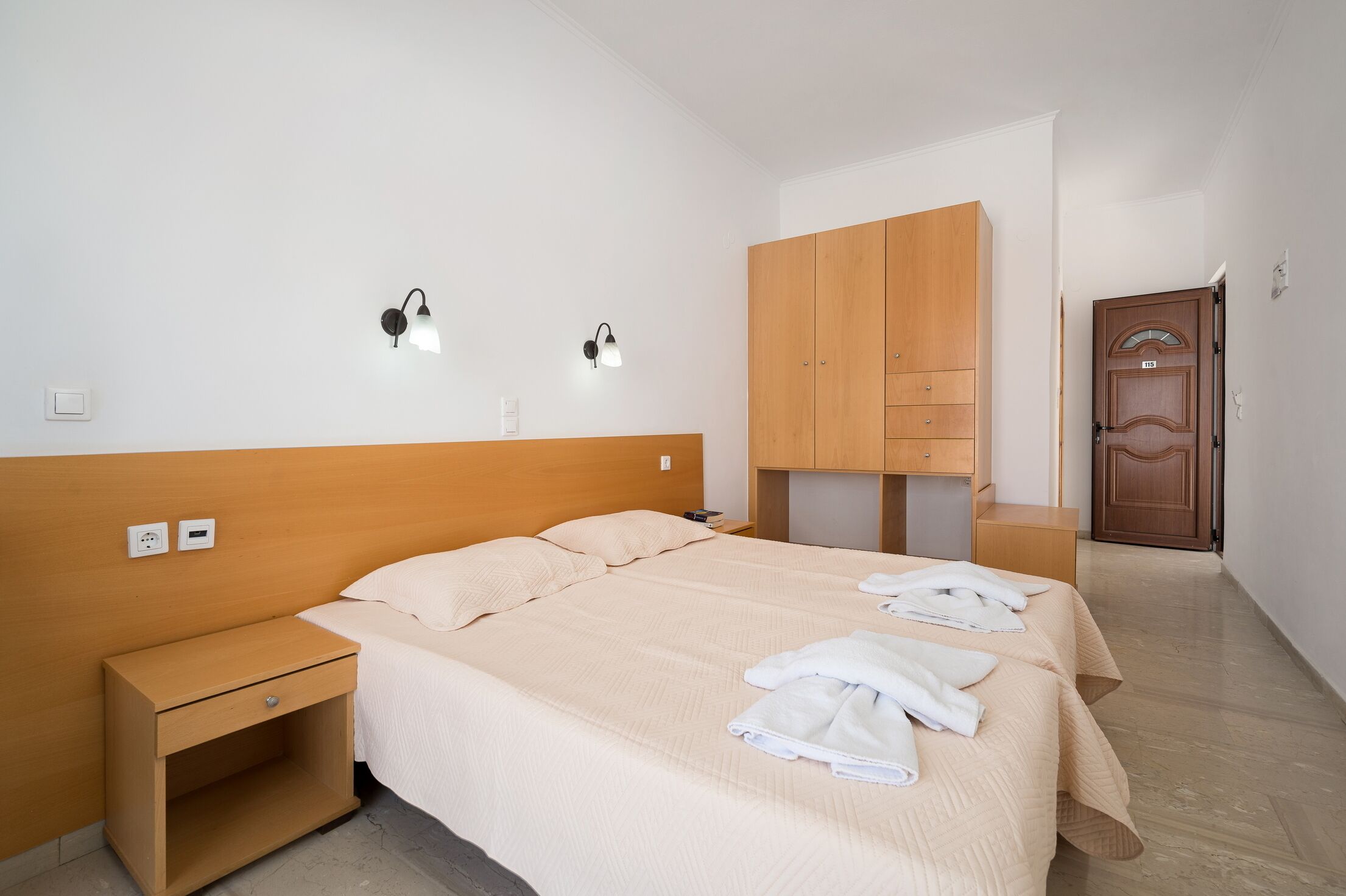 A hotel room with a bed, bedside table, wall-mounted lamp, wardrobe, and a door.