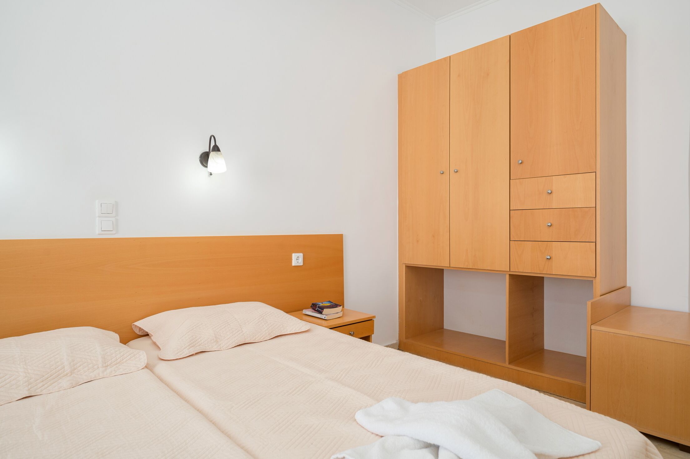 A bedroom with a bed, a nightstand, a wooden wardrobe, and a wall-mounted light fixture.
