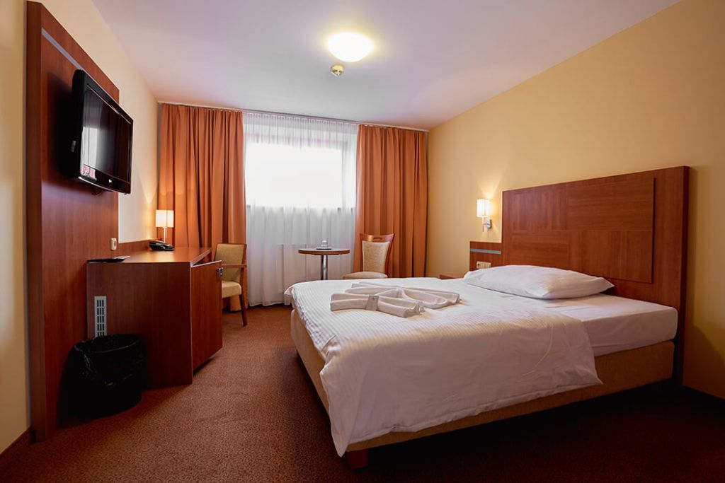 Habitació d'hotel amb un llit, una taula de nit, televisió, una cadira, una taula petita i una finestra amb cortines.