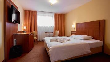 Habitació d'hotel amb un llit, una taula de nit, televisió, una cadira, una taula petita i una finestra amb cortines.