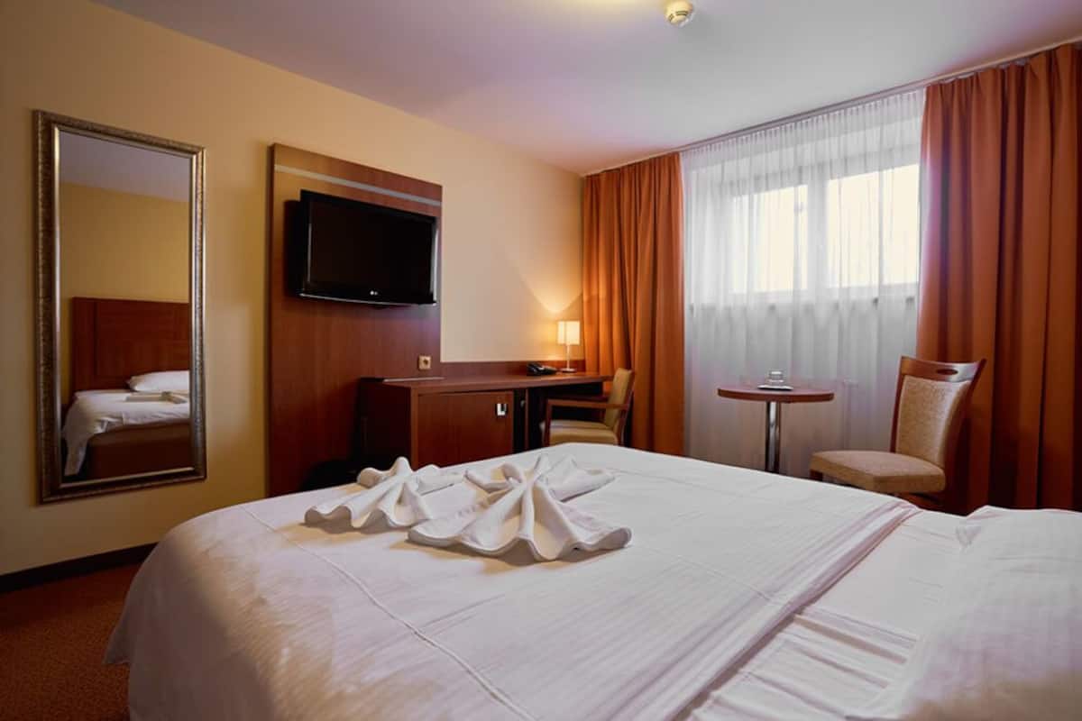 Quarto de hotel com uma cama grande, uma escrivaninha, uma cadeira, uma televisão e uma janela com cortinas.