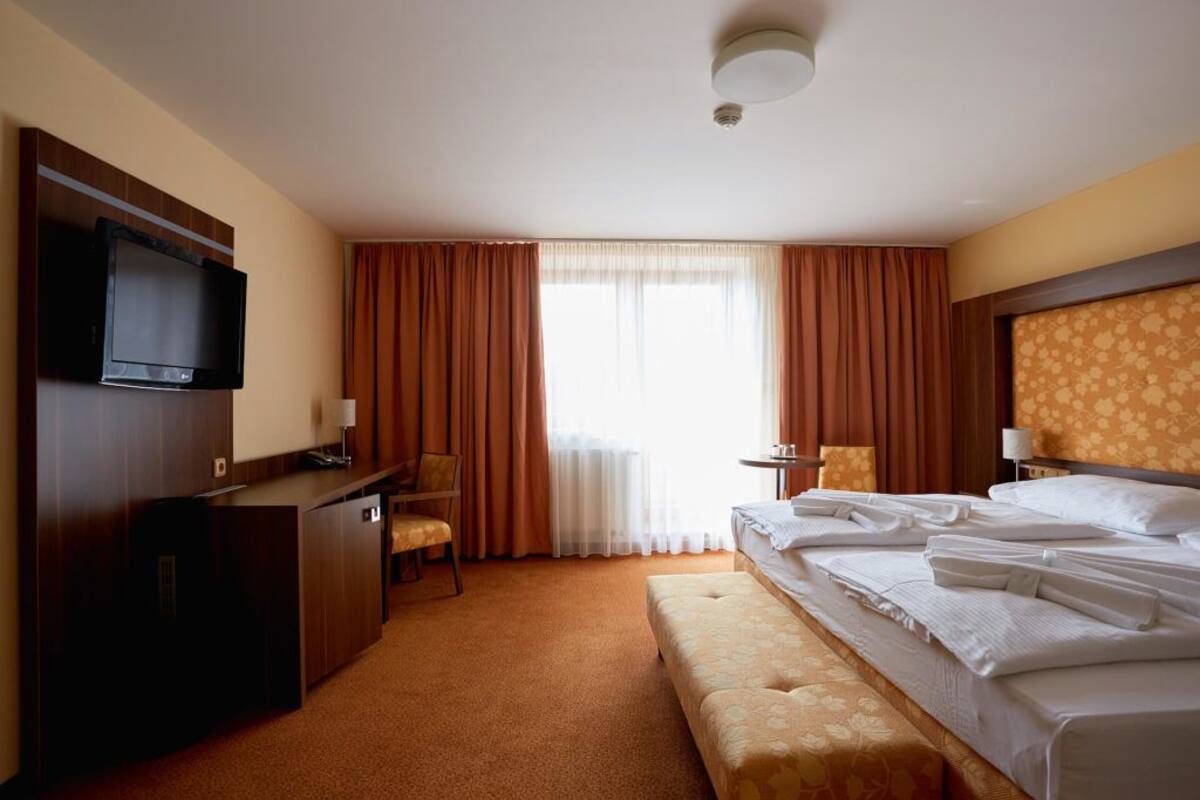 Quarto de hotel com cama, escrivaninha com cadeira, televisão e janela com cortinas.