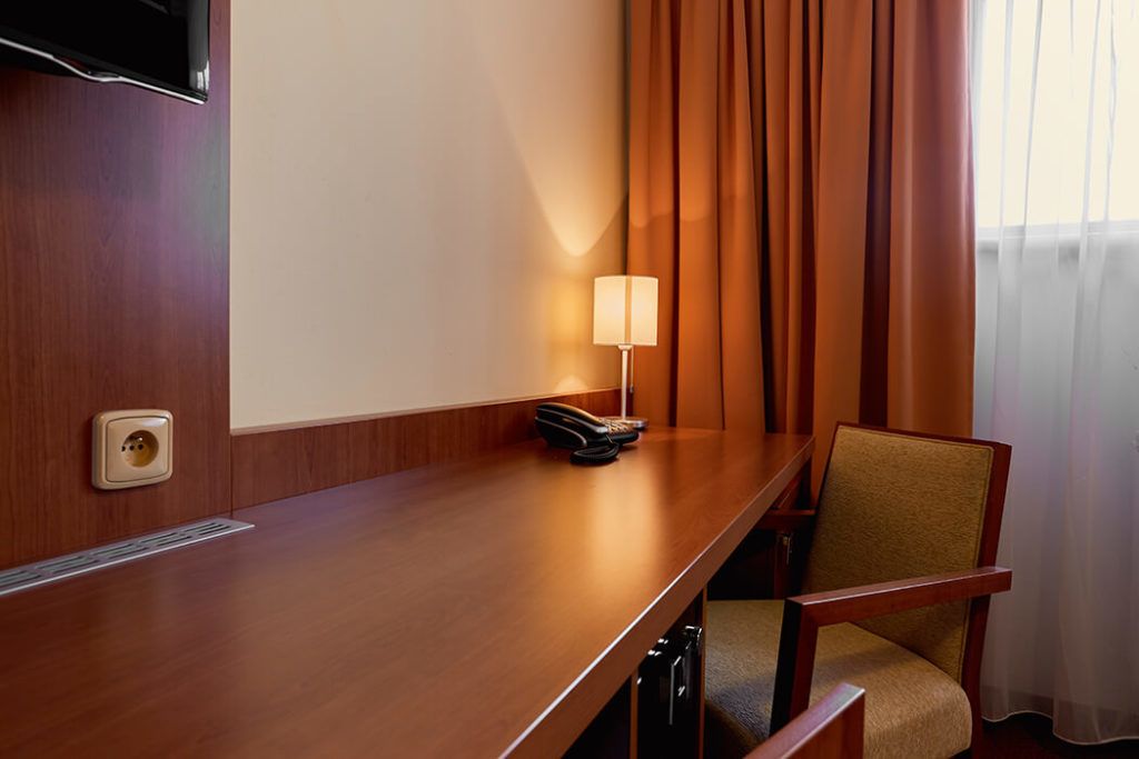 A hotel room with a wooden desk, a chair, a lamp, and a TV mounted on the wall.
