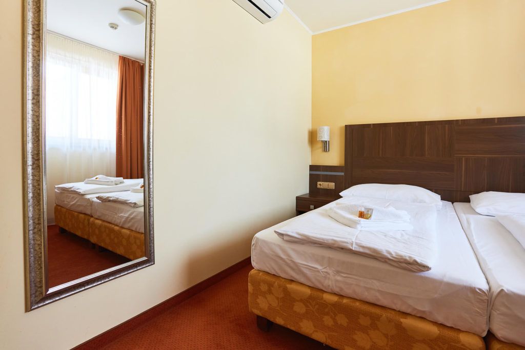 A hotel room with a large mirror, a bed with neatly made white linens, a wooden headboard, and a nightstand with a lamp.