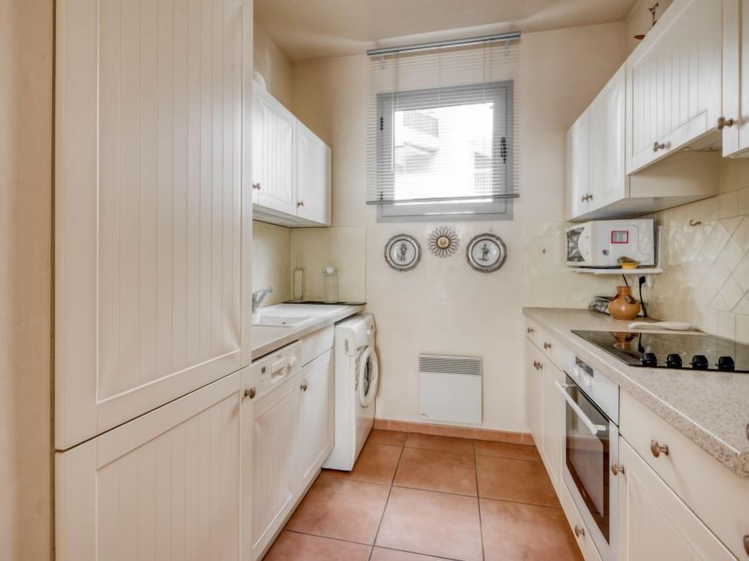 Une cuisine compacte avec des meubles blancs, un lave-linge encastré, un four à micro-ondes et une petite fenêtre avec des stores.