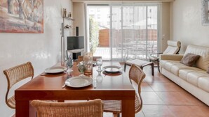 Un coin repas avec une table en bois dressée pour quatre personnes, entourée de chaises en osier. Un canapé et un téléviseur sont visibles au fond.