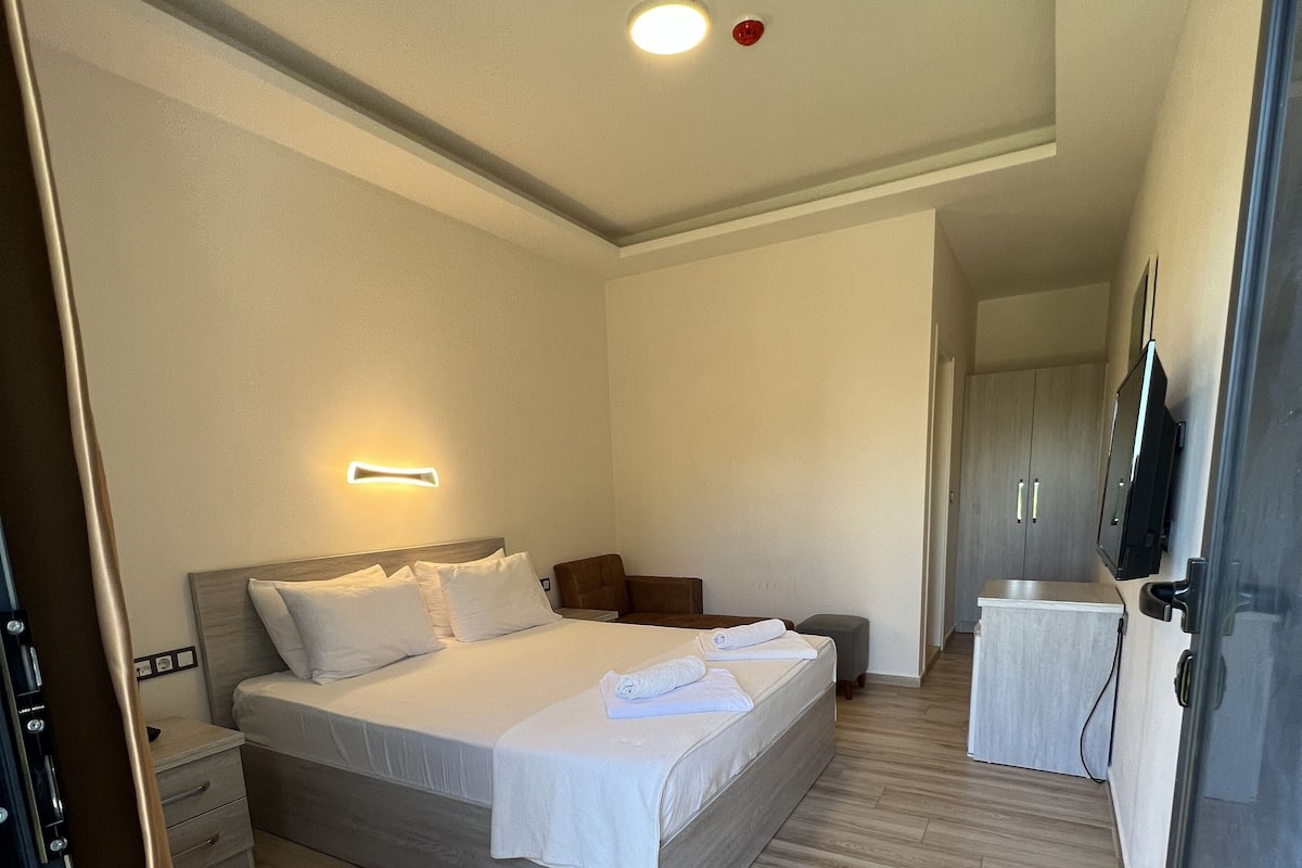 A modern hotel room with a large bed, bedside tables, a TV, and a wooden floor.