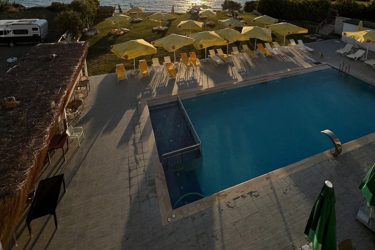 A pool area with sun loungers and umbrellas, a clear view of the sunset over the water, and a distant mountain range.