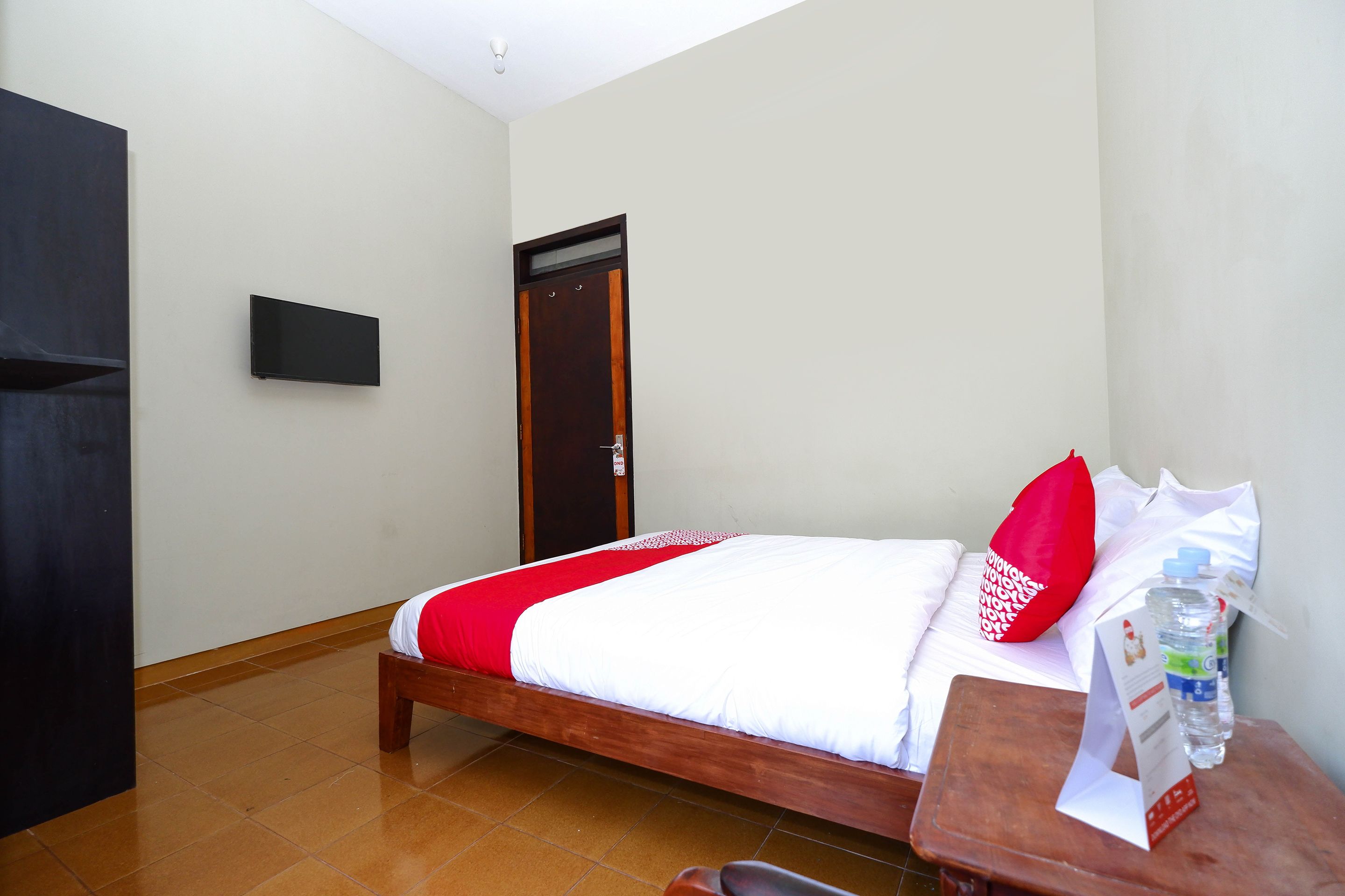 Een hotelkamer met een bed, een houten nachtkastje, een aan de muur bevestigde televisie en een deur.