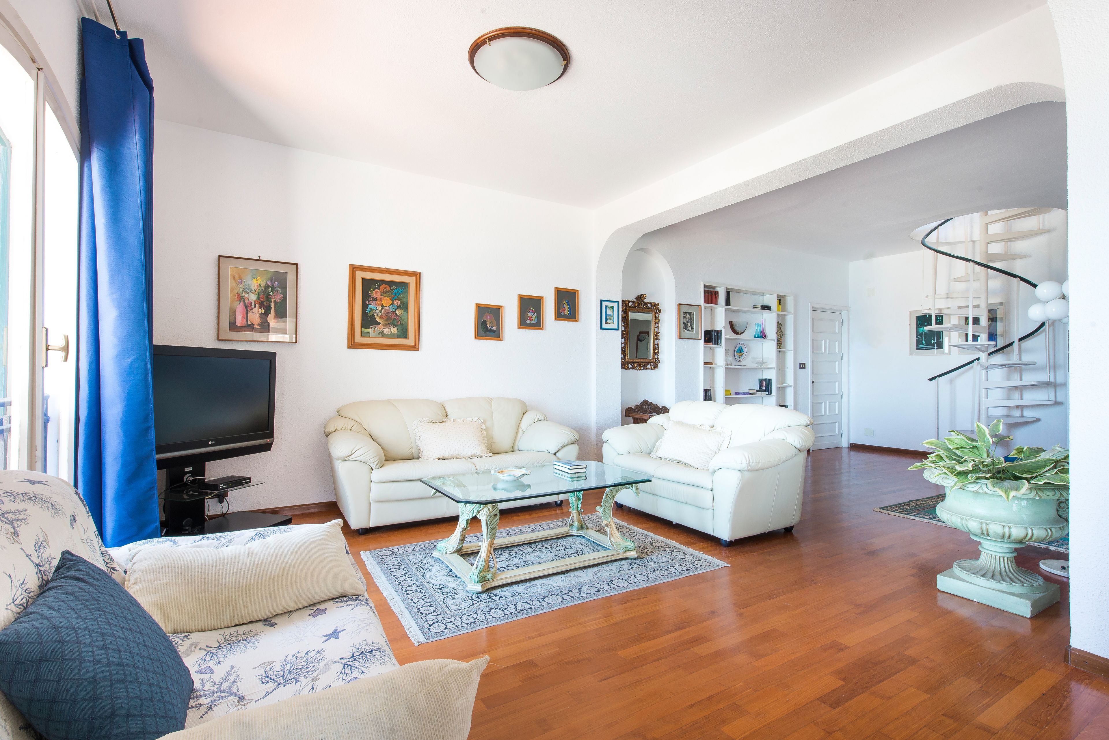A living room with a sofa, a glass coffee table, and a television. There are framed pictures on the wall and a potted plant.