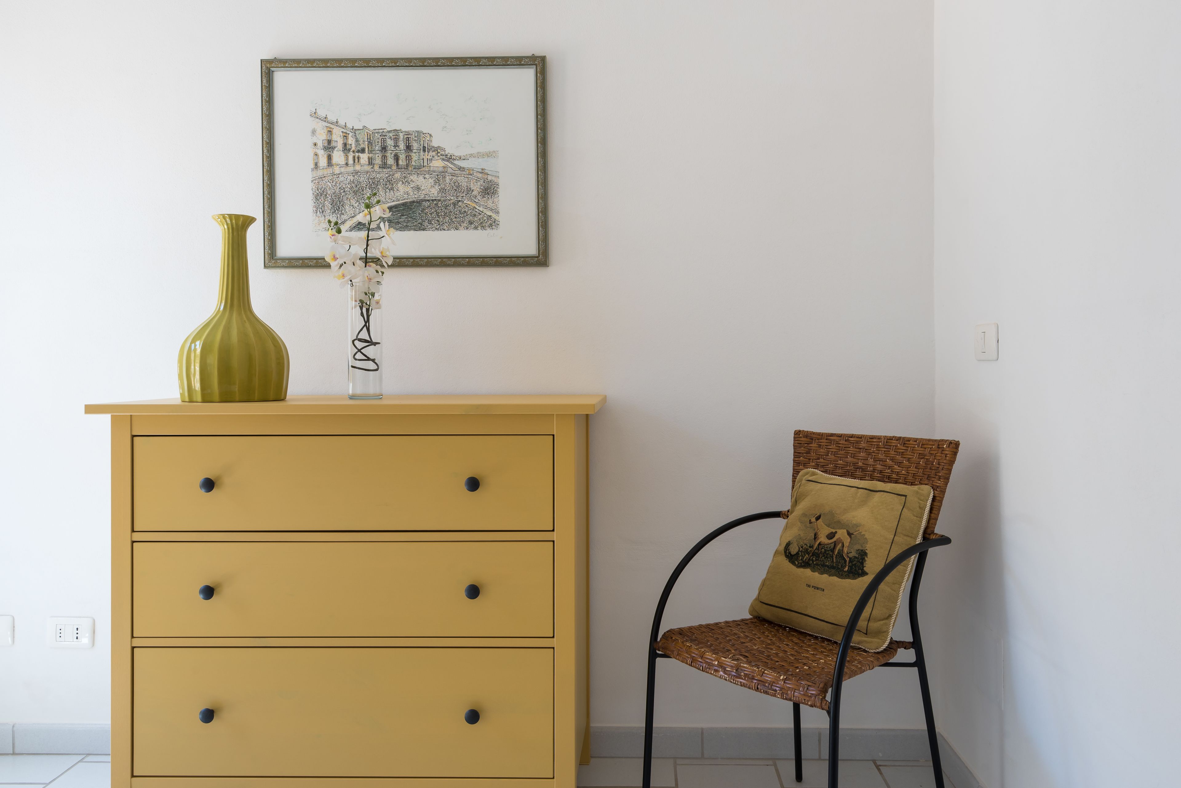 A room with a yellow dresser, a framed picture on the wall, a green vase with flowers, and a wicker chair with a cushion.