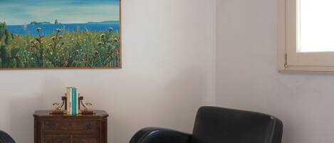 A room with a wooden ceiling, a painting of a landscape, a black leather chair, a wooden cabinet, and a window.