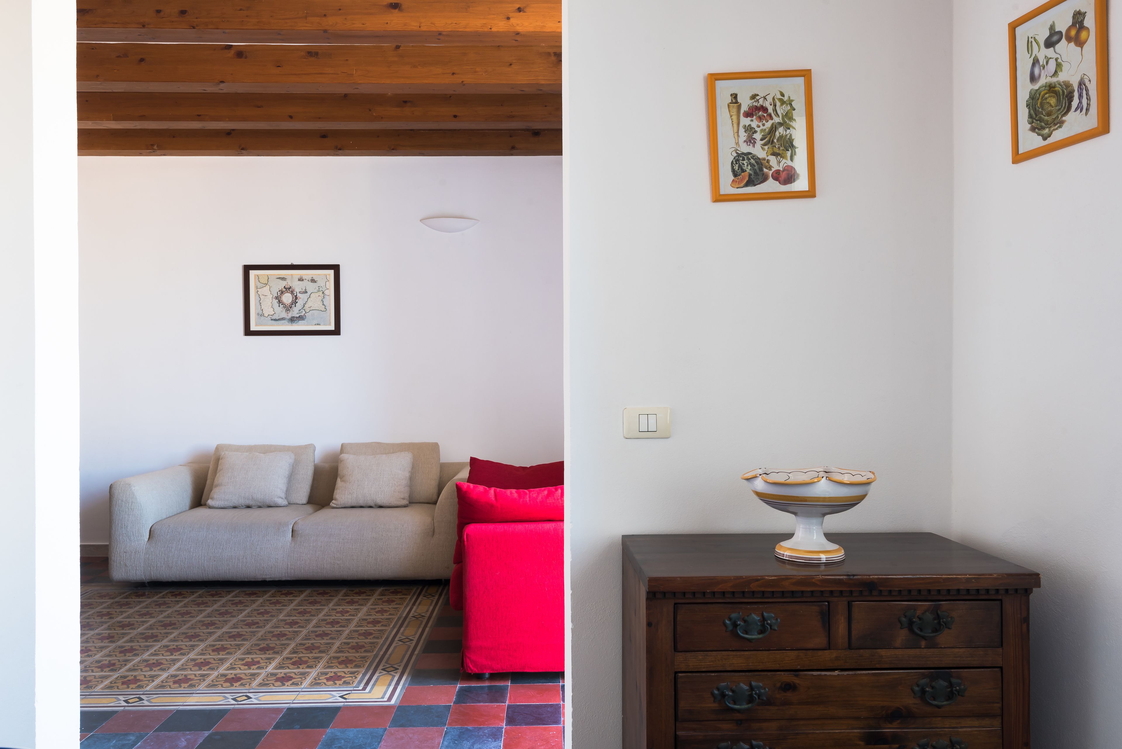 A room with a sofa, a red ottoman, a patterned rug, a wooden cabinet with a bowl, and framed botanical prints on the wall.