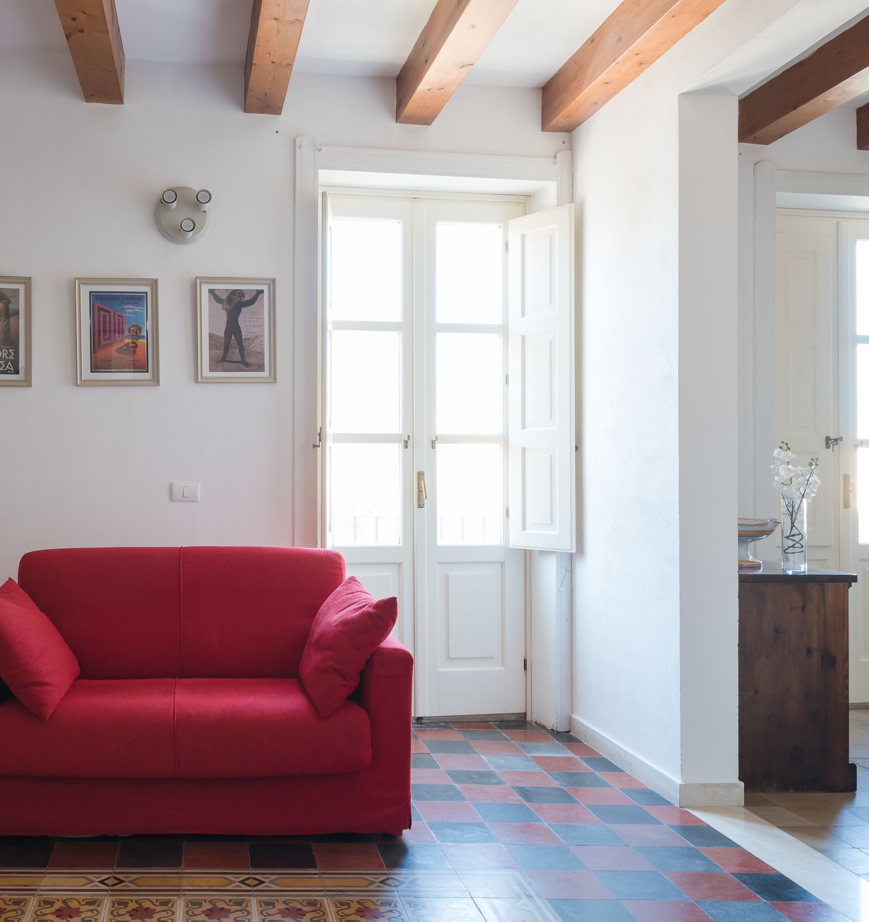 A red sofa in a room with a checkered floor, white walls, and wooden beams on the ceiling.
