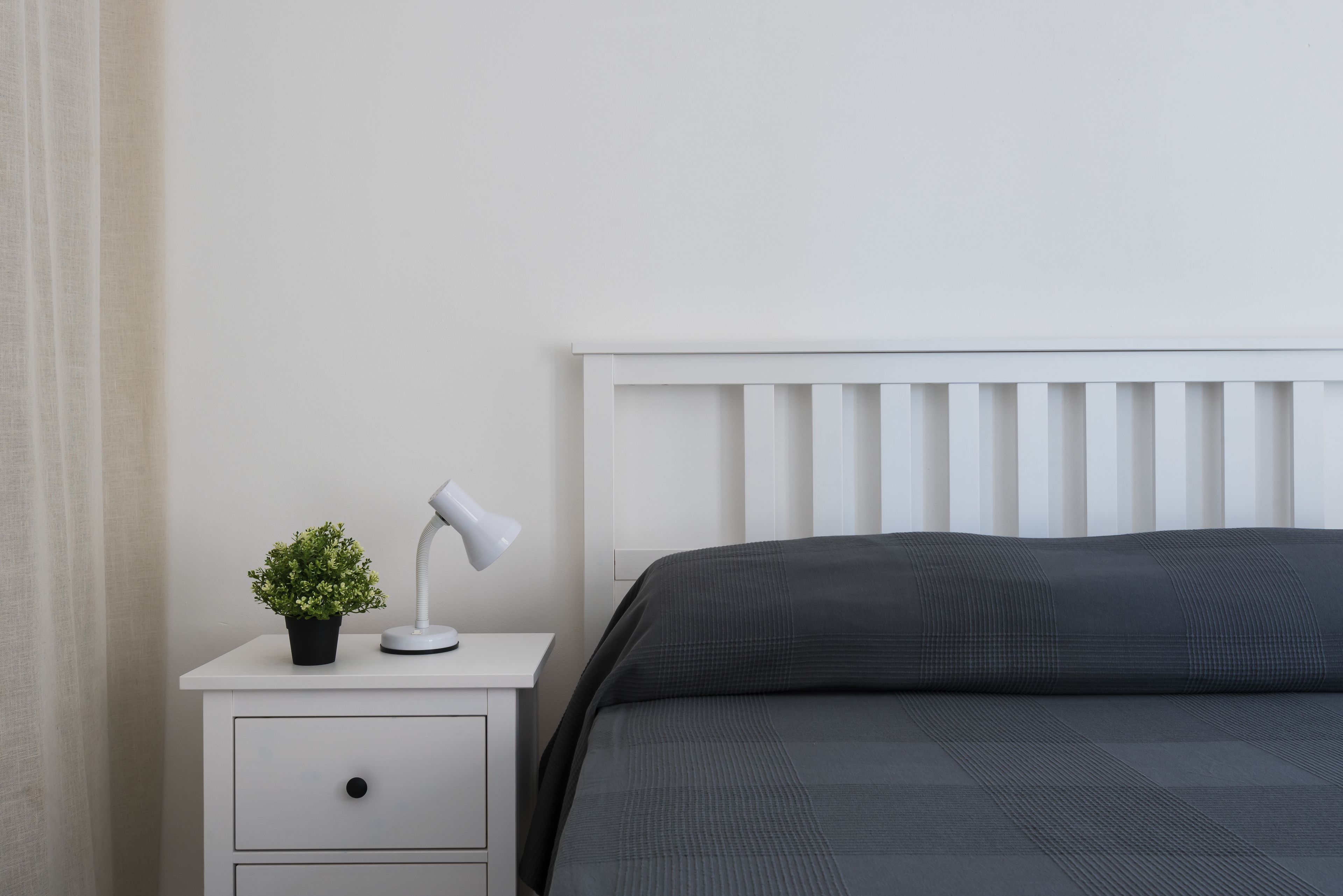 Ein ordentlich bezogenes Bett mit einer dunkelgrauen Tagesdecke, ein weißer Nachttisch mit Lampe und einer kleinen Pflanze sowie ein weißer Kopfteil.
