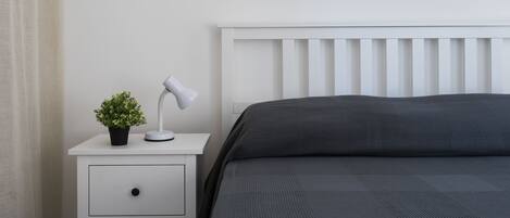 Ein ordentlich bezogenes Bett mit einer dunkelgrauen Tagesdecke, ein weißer Nachttisch mit Lampe und einer kleinen Pflanze sowie ein weißer Kopfteil.