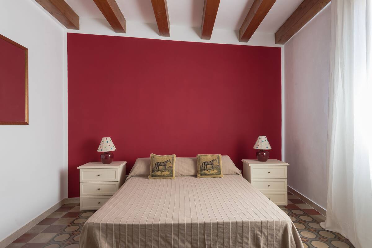 Une chambre avec un mur rouge, un lit avec une literie à rayures, deux tables de chevet avec des lampes et un sol à motifs.