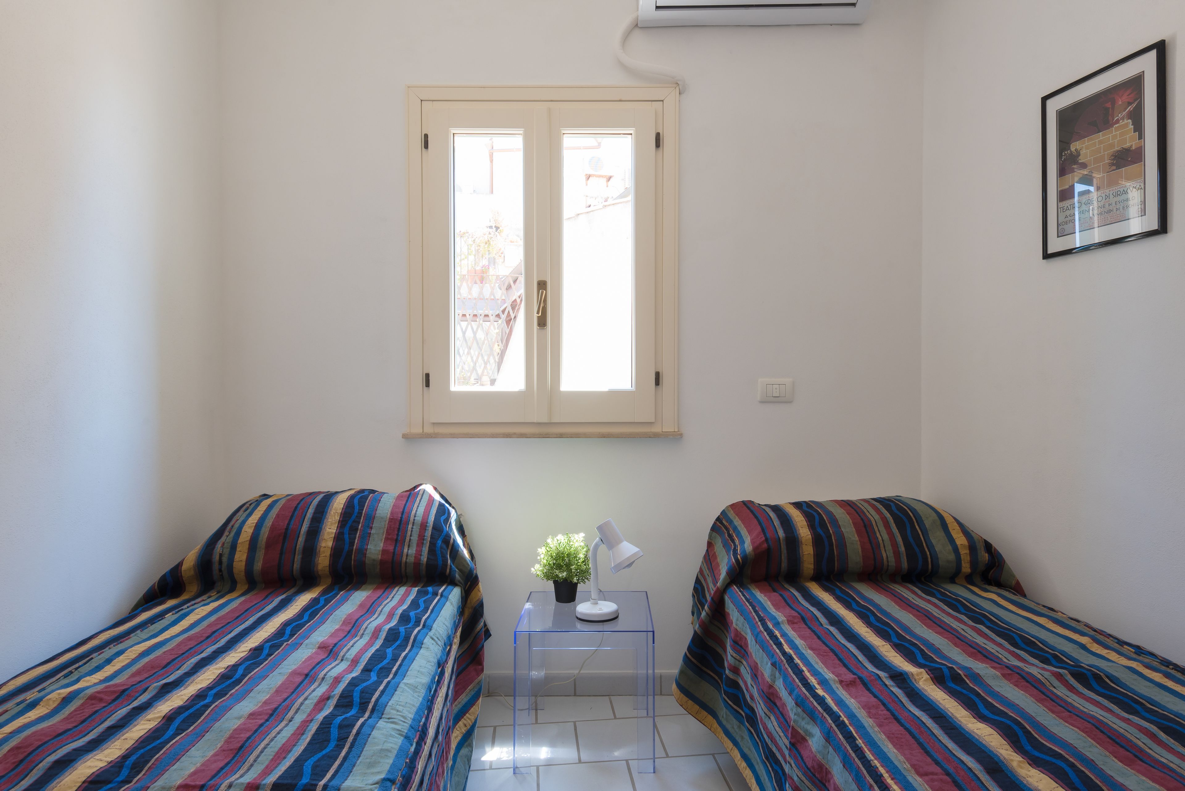 Quarto pequeno e bem iluminado com duas camas, uma mesinha com abajur e uma planta, e uma janela com vista para o exterior.