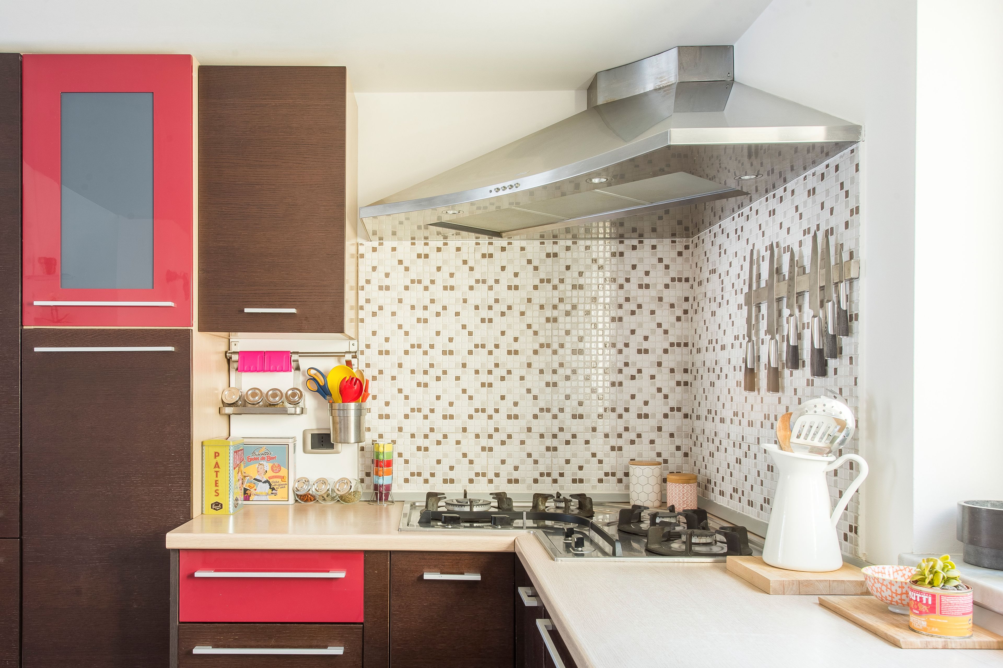 A kitchen with a gas stove, red and brown cabinets, a stainless steel range hood, and a tiled backsplash.