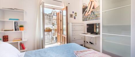 A bedroom with a bed, a wardrobe, a desk, and a balcony with a view of buildings.