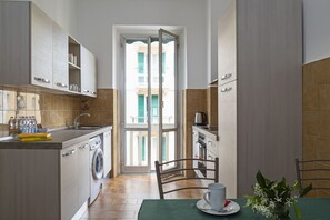 Une cuisine compacte équipée d’un lave-linge, d’une table à manger avec une tasse et une soucoupe, et donnant sur un balcon par une porte ouverte.