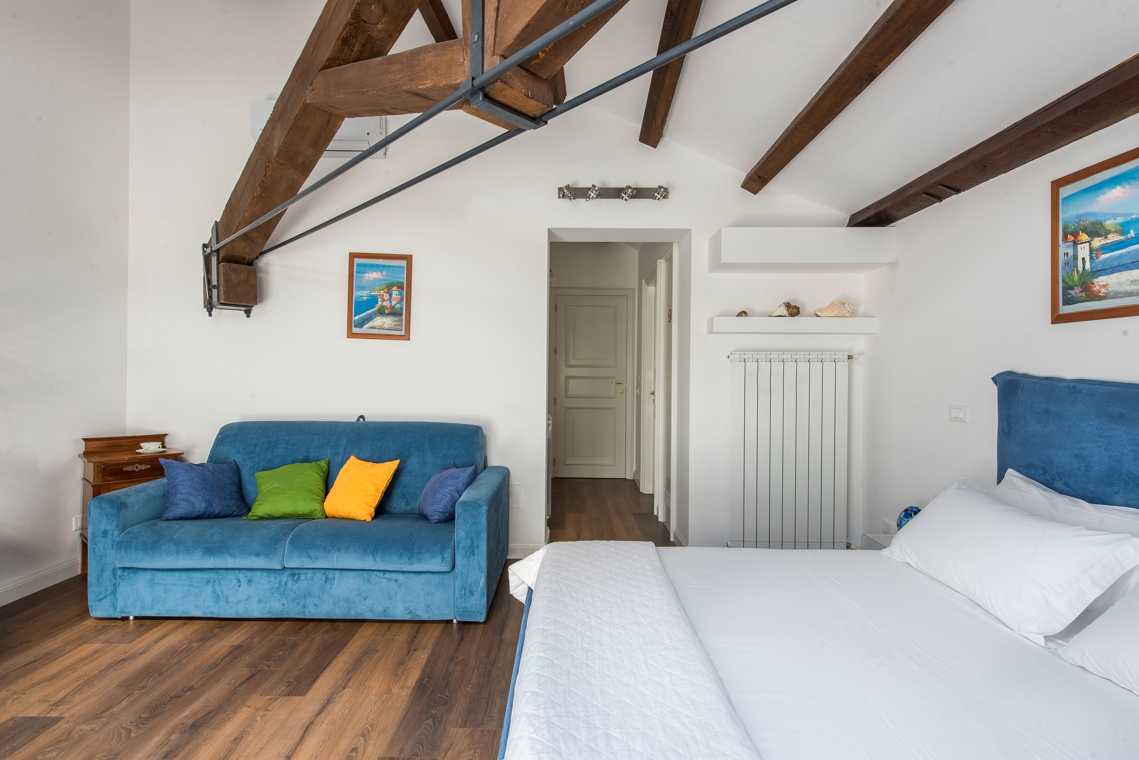 Ein Zimmer mit blauem Sofa, einem Bett, Holzbalken an der Decke, einem weißen Heizkörper und einer Tür zu einem anderen Raum.