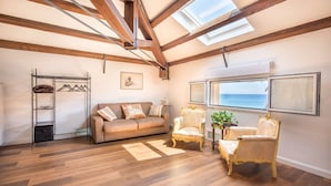 Une pièce spacieuse avec des poutres apparentes, un canapé, deux fauteuils décoratifs, un porte-manteau en métal et une grande fenêtre donnant sur la mer.