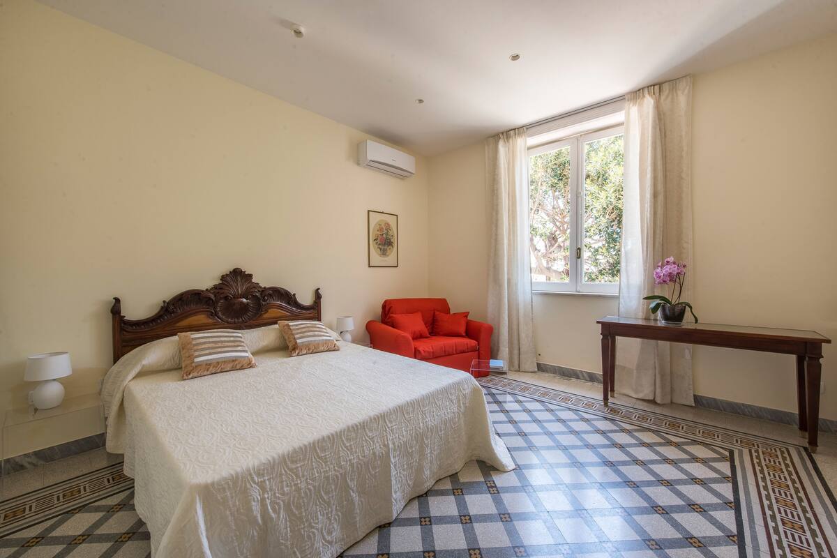 Een slaapkamer met een groot bed, een rode fauteuil, een klein tafeltje en een raam met gordijnen.