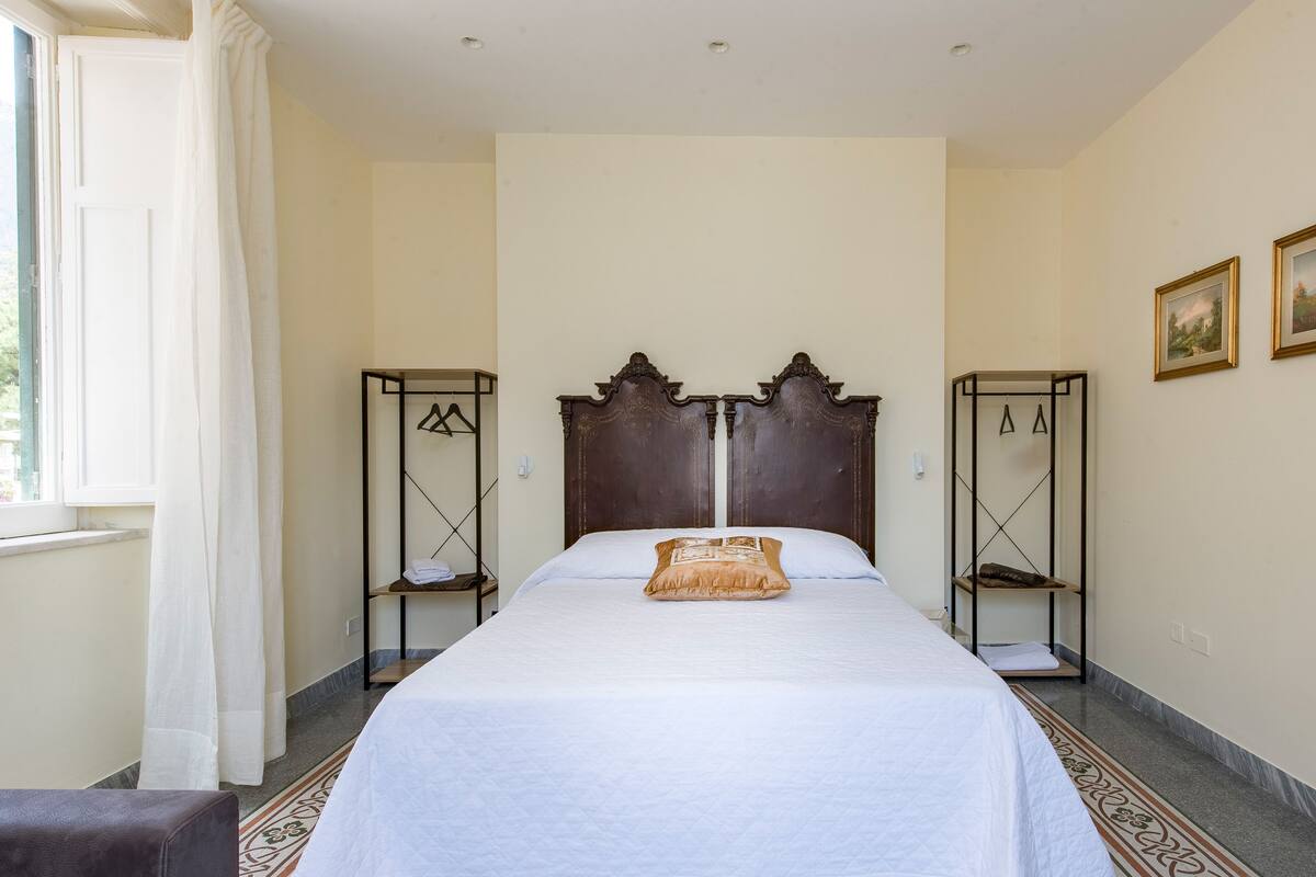 Een hotelkamer met een groot bed, een houten hoofdeinde, een klein plankje en een raam met gordijnen.