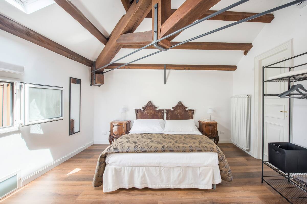 Een slaapkamer met een houten bedframe, een groot raam, een radiator en een zwarte opbergunit.