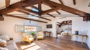 Un vaste salon doté d’un plancher en bois, d’un plafond haut avec des poutres apparentes, d’un canapé, de deux fauteuils, d’un téléviseur et d’un coin repas à l’arrière-plan.