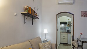 A living room with a sofa, a lamp, and a bookshelf. There is a kitchen visible through an archway.