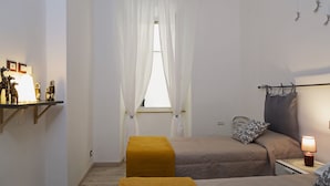 A bedroom with two beds, a window with sheer curtains, a bedside table with a lamp, and a shelf with decorative items.