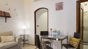 A compact living room with a sofa, a small side table with a lamp, a dining table with chairs, a large mirror, and a clock on the wall.