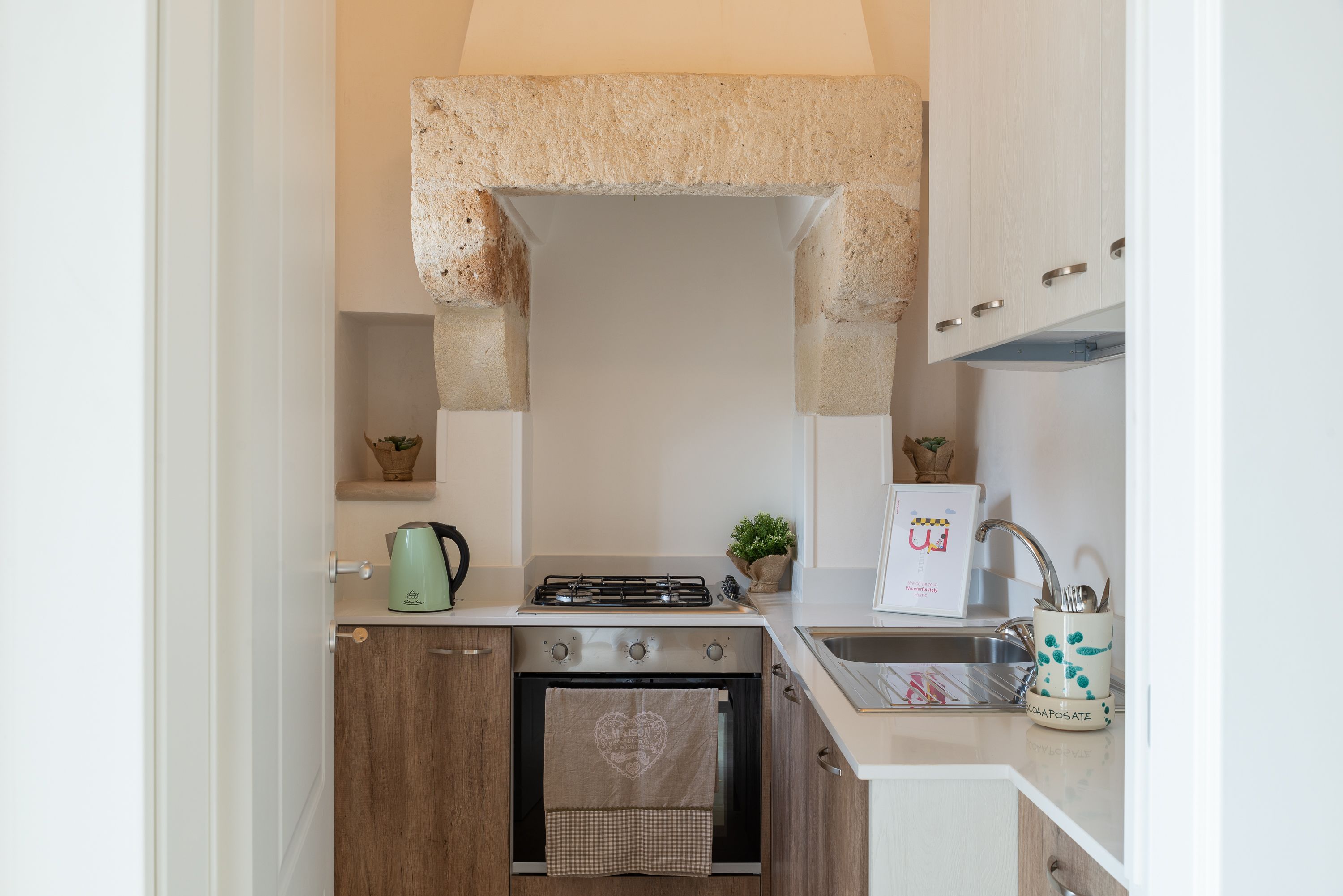 A compact kitchen with a gas stove, oven, and sink. There is a small countertop with a kettle and a plant.