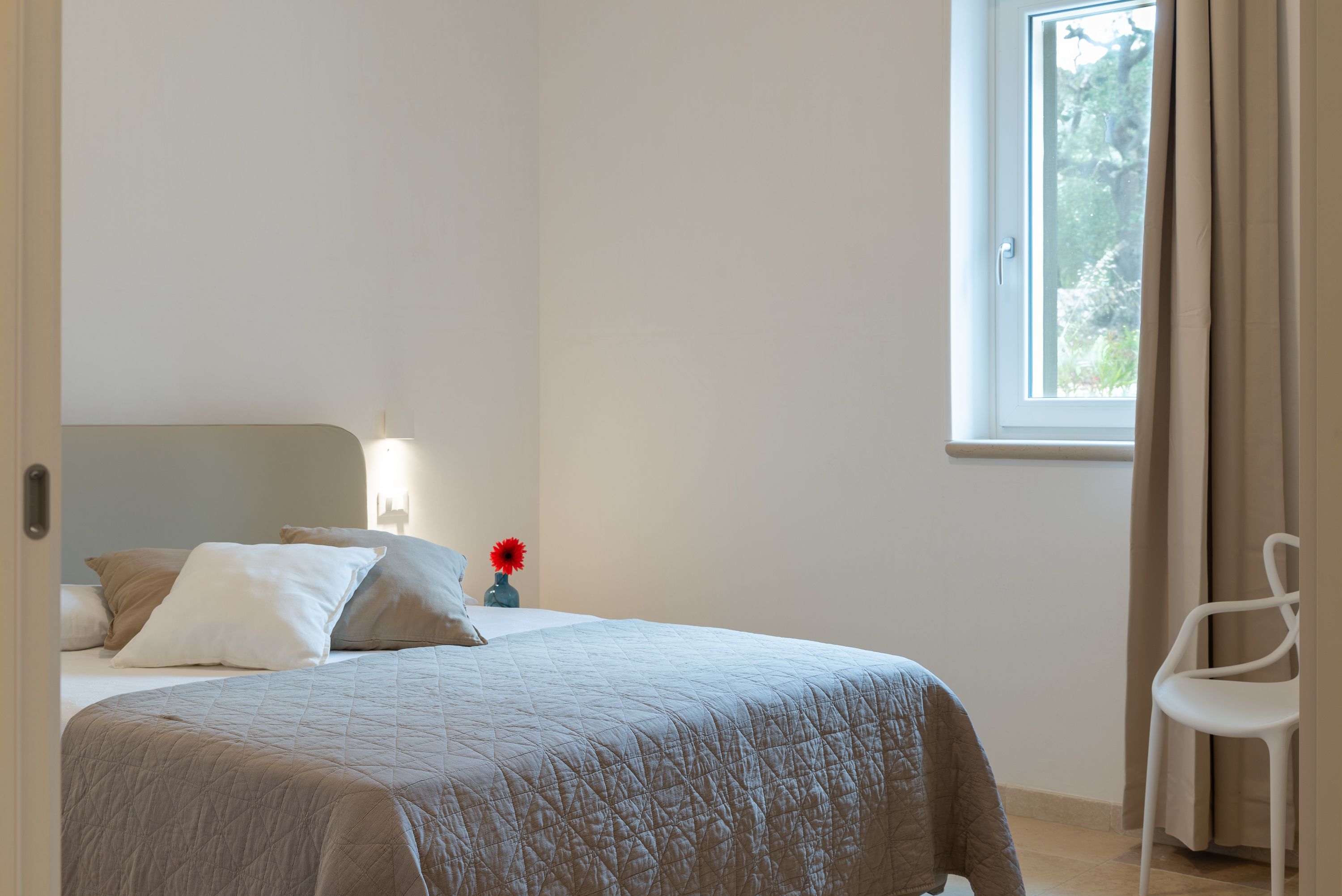 A bedroom with a bed, a nightstand with a vase, a chair, and a window with curtains.