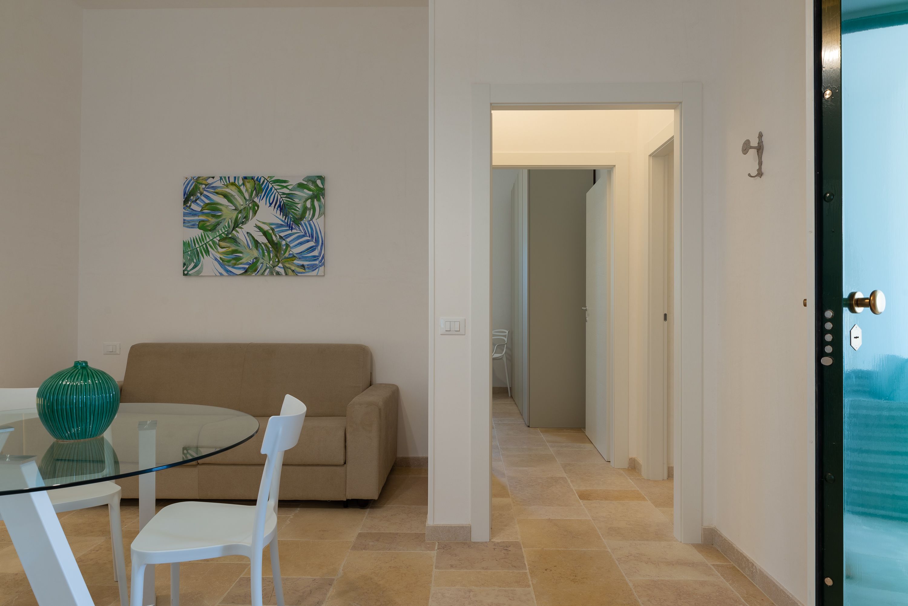 A living room with a glass table, white chairs, a beige sofa, and a green vase. There is a painting on the wall and a door leading to another room.