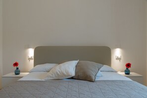 Een net opgemaakt bed met een gewatteerde spreidek, twee nachtkastjes met lampen, en twee vazen met rode bloemen.
