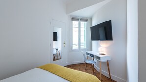 A bedroom with a bed, a desk, a chair, a TV, and a window.