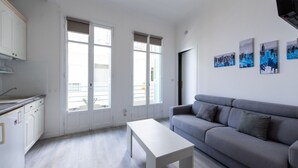 A modern living room with a grey sofa, a white coffee table, and a kitchen area with white cabinets and a window.