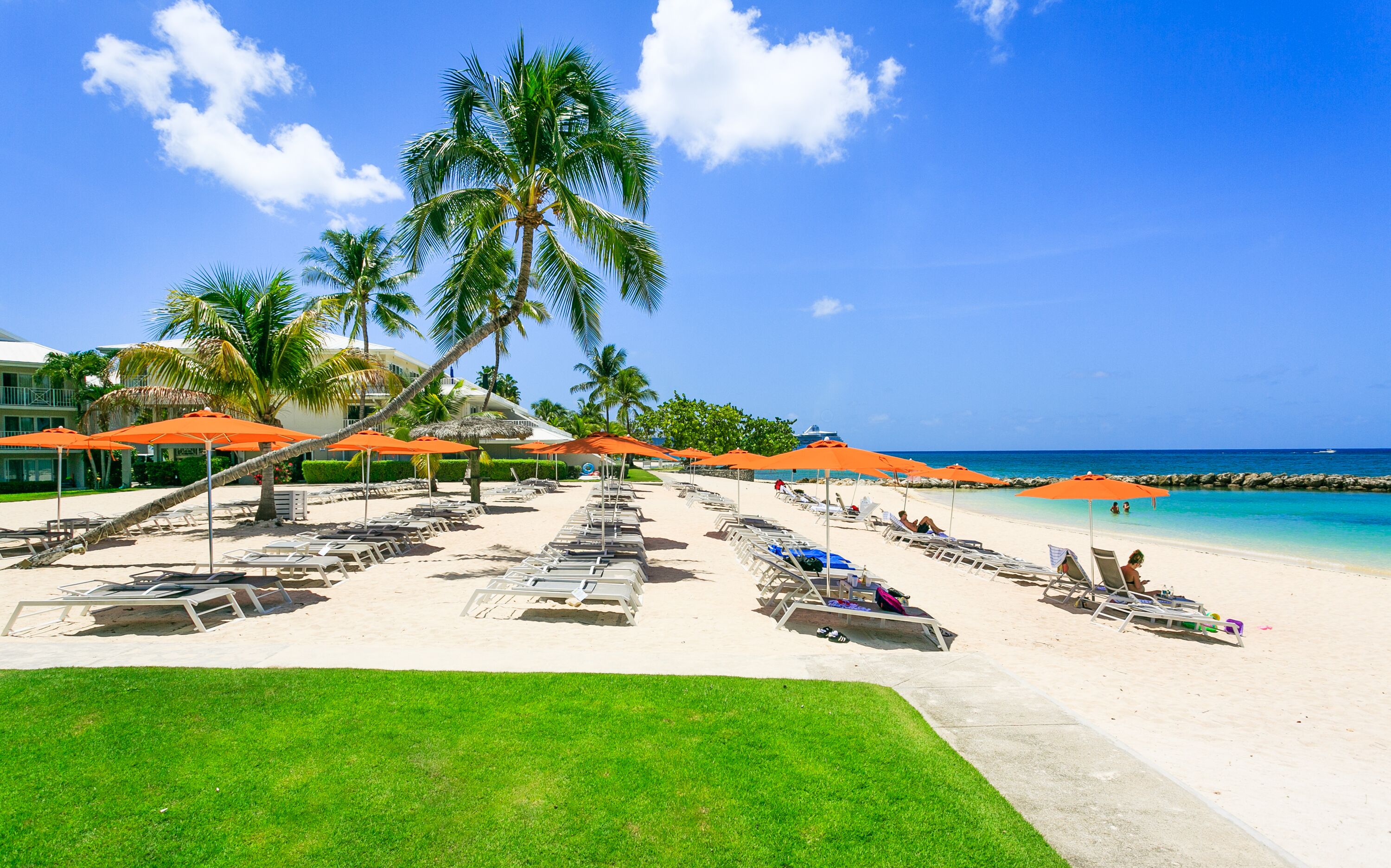 A beach resort with rows of sun loungers and orange umbrellas, palm trees, and a clear blue sky.